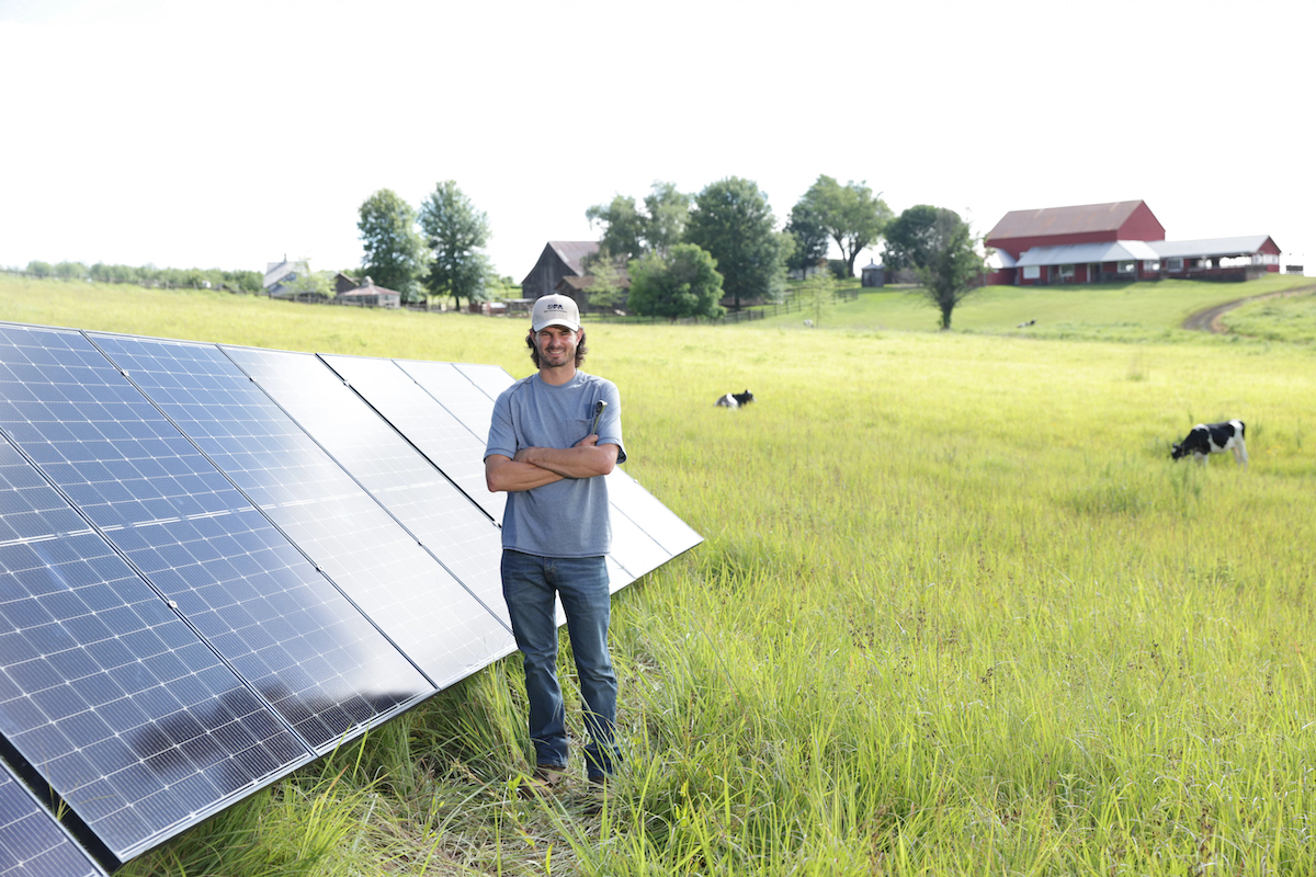 sustainability A man walks across a grassy field with cows grazing nearby. Behind him are two solar-powered wind turbines, each equipped with solar panels and red turbines, set against a clear blue sky.