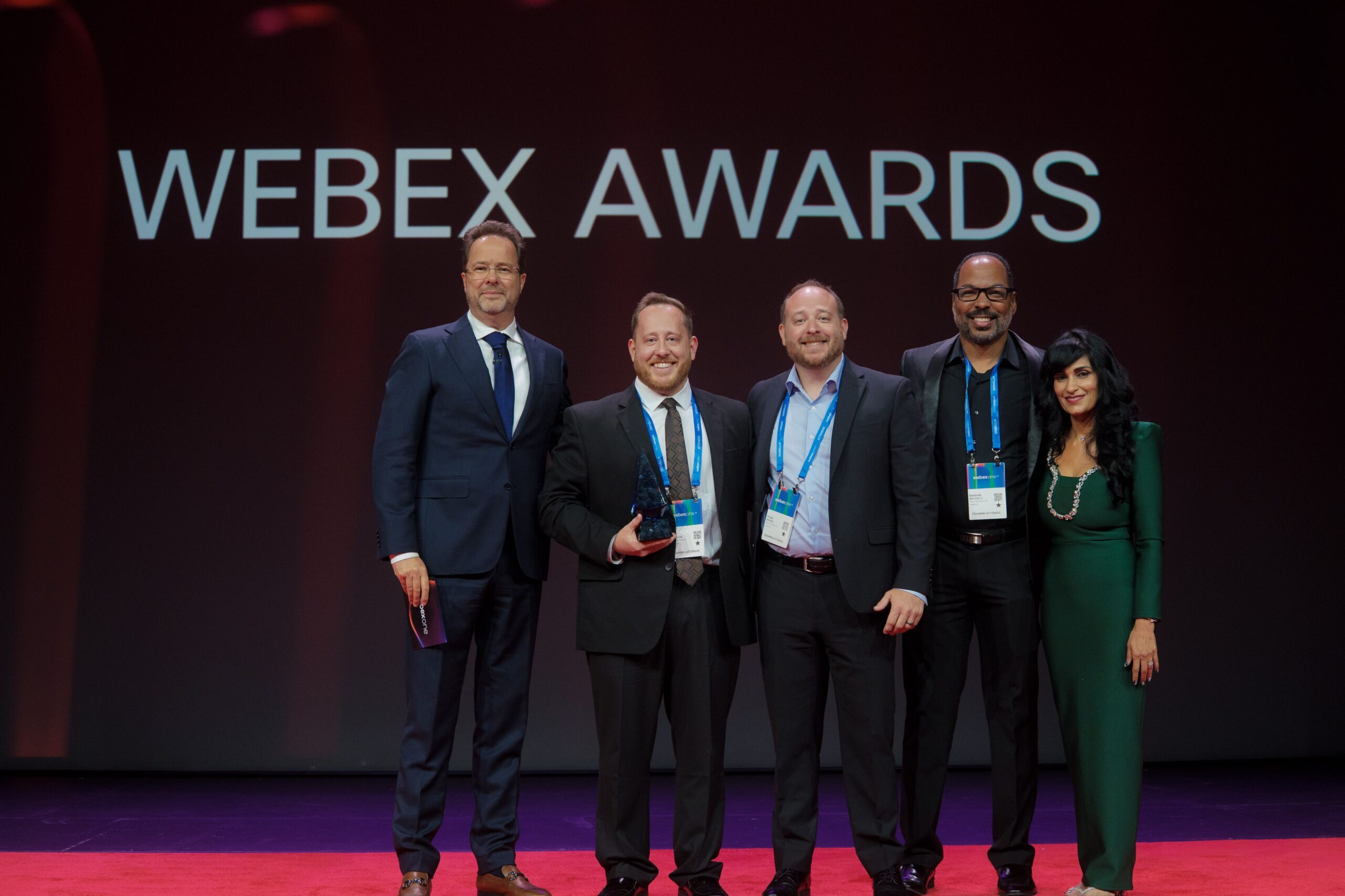 Four people in business attire stand posing indoors; the person second from the left holds a plaque. A decorative wall with bottle-shaped designs is in the background.